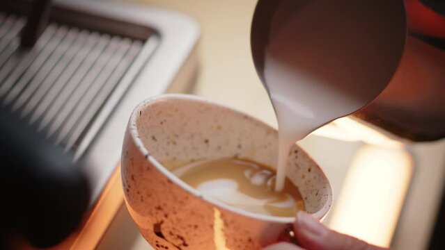 Closeup shot of pouring foamed milk into a beautiful cup of coffee. Barista making cappuccino in a morning cafe. Warm lighting