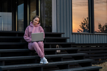 Obraz premium Woman dressed in a pink suit sitting on the stairs of her house in the sunlight on a summer evening and using a laptop. 