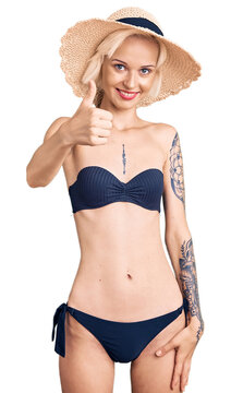 Young blonde woman with tattoo wearing bikini and summer hat doing happy thumbs up gesture with hand. approving expression looking at the camera showing success.