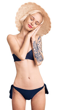 Young blonde woman with tattoo wearing bikini and summer hat sleeping tired dreaming and posing with hands together while smiling with closed eyes.