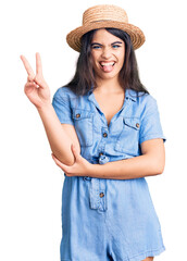 Brunette teenager girl wearing summer hat smiling with happy face winking at the camera doing victory sign. number two.