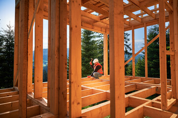 Carpenter constructing two-story wooden frame house near forest. Man hammering nails into the structure, wearing protective helmet and construction vest. Concept of modern ecological construction.
