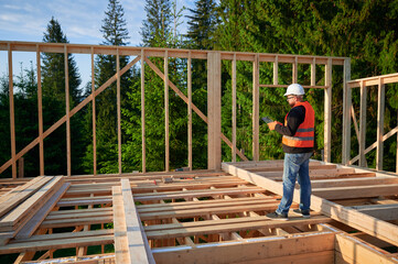 Carpenter constructing wooden two-story frame house near the forest. Bearded man in jumpsuit and protective helmet examining construction plan. Concept of modern ecological construction.