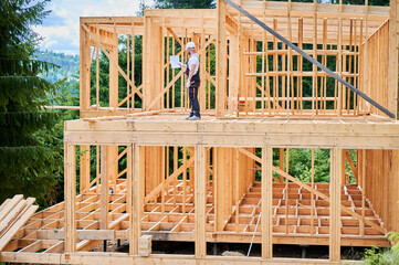 Inspector examining construction plan of wooden two-story building. Man, dressed in jumpsuit and...