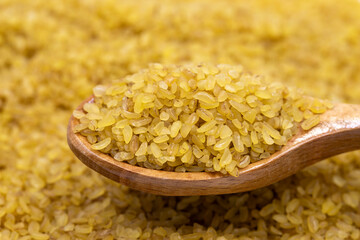 Raw bulgur wheat on wooden spoon, legumes as background. close-up uncooked dry bulgur wheat