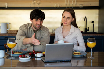 Happy young couple husband and wife use a laptop while looking at the screen, pay bills online in applications, calculate an investment mortgage payment on a website while sitting at the dining table