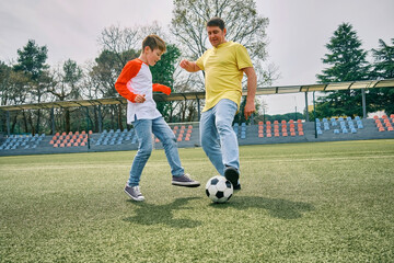 Obraz premium Father and Son play football on stadium outdoors, Happy family bonding, fun, players in soccer in dynamic action playing in sunny day, holidays time. 