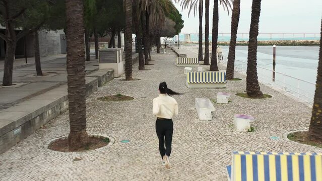Aerial video of Caucasian woman running waterfront public park. Active runner alone in sportswear exercising outdoor. Healthy lifestyle concept. Back view