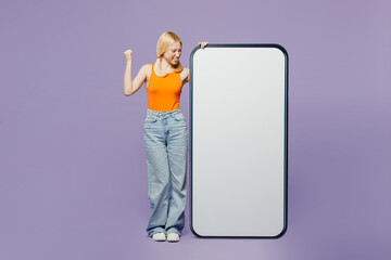 Full body young blonde woman she wear orange tank shirt casual clothes big huge blank screen mobile cell phone smartphone with area do winer gesture isolated on plain pastel light purple background.