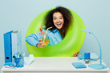 Young fun employee business woman wear casual shirt sit work at white office desk dream about free time vacations hold inflatable ring drink cocktail isolated on plain pastel blue background studio.