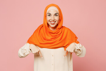 Fototapeta premium Young arabian asian muslim woman wear orange abaya hijab point index finger down on area workspace isolated on plain light pink background studio portrait. Uae middle eastern islam religious concept.