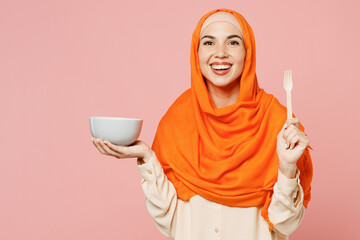 Young happy fun arabian asian muslim woman wear orange abaya hijab hold in hand fork bowl with meal isolated on plain light pink background studio portrait. Uae middle eastern islam religious concept