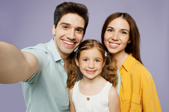 Close up young happy parents mom dad with child kid daughter girl 6 year old in blue yellow casual clothes do selfie shot pov mobile cell phone isolated on plain purple background Family day concept