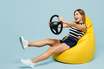 Full body young woman she wear striped tank shirt casual clothes sit in bag chair hold steering wheel driving car isolated on plain pastel light blue cyan background studio portrait Lifestyle concept - obrazy, fototapety, plakaty