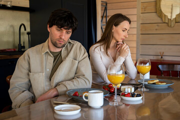 Stressed young married couple sits separately on opposite sides of the dining table, ignoring each other after a quarrel. Offended spouses do not talk, communicate, feeling depressed and disappointed