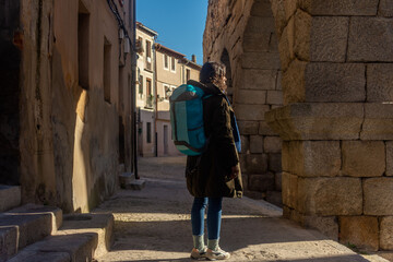 Woman wearing a coat, carrying a backpack, standing in front of an ancient stone wall with arches
