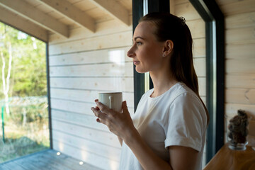 Woman with coffee mug standing in the living room looking out of terrace door. 