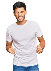 Fototapeta premium Young handsome man wearing casual white tshirt very happy and excited doing winner gesture with arms raised, smiling and screaming for success. celebration concept.