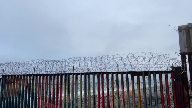 Camera follows a flock of birds as they gently fly over the barbed-wire-topped border wall that separates the United Stated and Mexico. Filmed on a cloudy day in the Tijuana coast (Mexico).