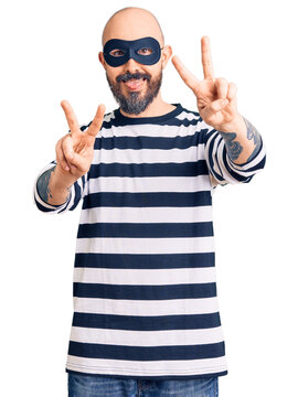 Young handsome man wearing burglar mask smiling with tongue out showing fingers of both hands doing victory sign. number two.