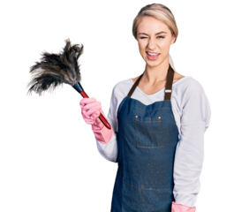 Beautiful young blonde woman wearing apron holding cleaning duster winking looking at the camera with sexy expression, cheerful and happy face.