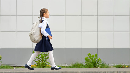 Schoolgirl with long hair carrying white backpack and blue book goes to modern school. Elementary...