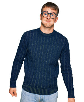 Young blond man wearing casual clothes and glasses puffing cheeks with funny face. mouth inflated with air, crazy expression.