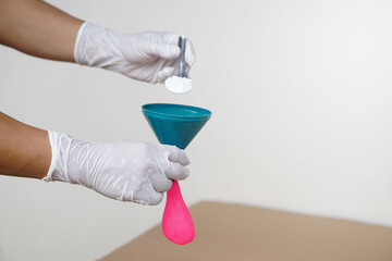 Closeup hands wears gloves holds funnel and flat pink balloon, put spoon of baking soda powder to...