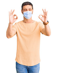 Handsome young man with bear wearing medical mask for coronavirus relax and smiling with eyes closed doing meditation gesture with fingers. yoga concept.