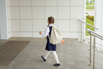 Brown-haired girl with white backpack entering primary school building. Junior schoolgirl in uniform enjoys walking alone to modern school