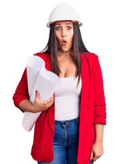 Young beautiful brunette woman wearing hardhat holding architect blueprint scared and amazed with open mouth for surprise, disbelief face