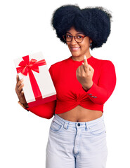 Young african american girl holding gift beckoning come here gesture with hand inviting welcoming happy and smiling