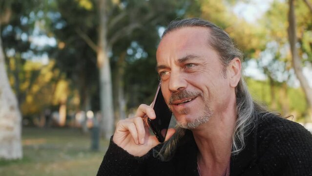 Handsome grey-haired mature man talking using smartphone with friends. Happy smiling middle aged hispanic person on the street.