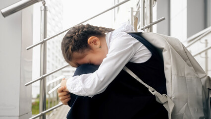 Brown-haired schoolgirl sitting on empty stairs cries remembering bullying moments. Girl with long braids afraid to attend school and see bullies - obrazy, fototapety, plakaty