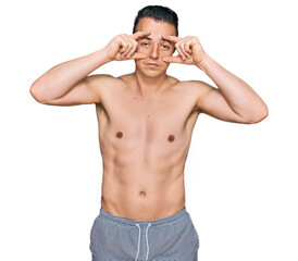 Fototapeta premium Handsome young man wearing swimwear shirtless trying to open eyes with fingers, sleepy and tired for morning fatigue