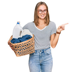 Beautiful blonde woman holding laundry basket and detergent bottle smiling happy pointing with hand and finger to the side