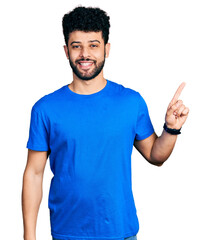 Fototapeta premium Young arab man with beard wearing casual blue t shirt with a big smile on face, pointing with hand finger to the side looking at the camera.