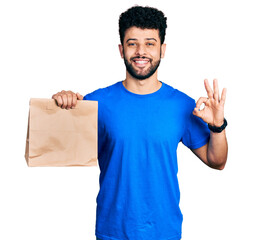 Young arab man with beard holding take away paper bag doing ok sign with fingers, smiling friendly gesturing excellent symbol