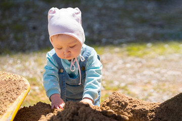 Toddler girl enjoys playing and collecting sand from playground. Daughter wearing pink hat spends daytime on playground illuminated by sun, close-up