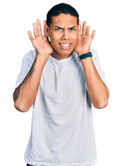 Fototapeta premium Young hispanic man wearing casual white t shirt trying to hear both hands on ear gesture, curious for gossip. hearing problem, deaf