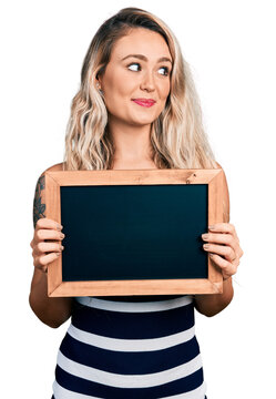 Young blonde woman holding blackboard smiling looking to the side and staring away thinking.