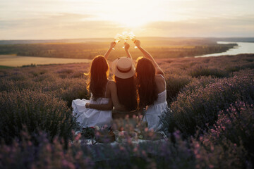 Young female friends having fun, raising glasses with wine and enjoy beautiful sunset at summer picnic in lavender field.	
