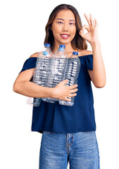 Young beautiful chinese girl holding recycling plastic bottles doing ok sign with fingers, smiling friendly gesturing excellent symbol