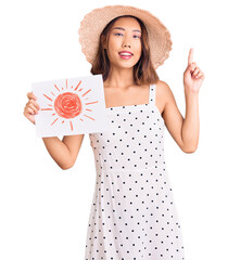 Young beautiful chinese girl wearing summer hat holding sun draw surprised with an idea or question pointing finger with happy face, number one