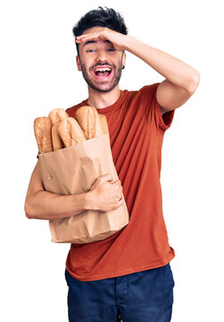 Young hispanic man holding paper bag with bread stressed and frustrated with hand on head, surprised and angry face
