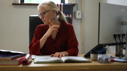 Closeup of happy woman teacher sitting at desk in empty school classroom desk grading papers working on assignments. US American flag in the background.
