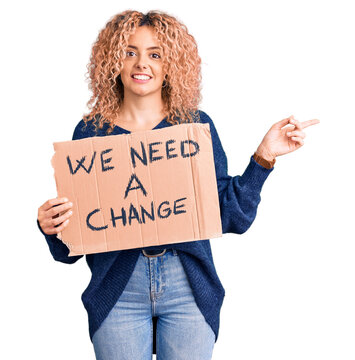 Young blonde woman with curly hair holding we need a change banner smiling happy pointing with hand and finger to the side