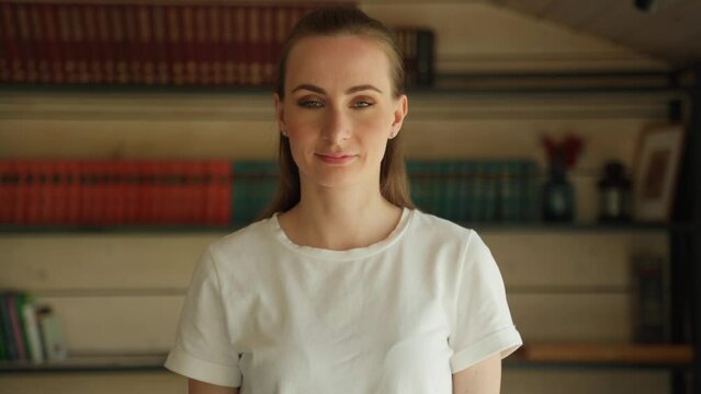 Portrait of a confident, happy, attractive student standing against the background of bookshelves in the campus library, looking at the camera, smiling amiably.