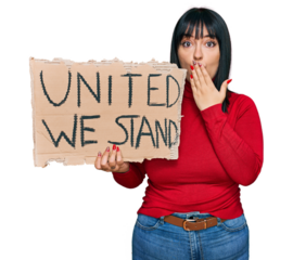 Young hispanic woman holding united we stand banner covering mouth with hand, shocked and afraid for mistake. surprised expression