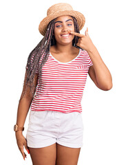 Obraz na płótnie Canvas Young african american woman with braids wearing summer hat pointing with hand finger to face and nose, smiling cheerful. beauty concept